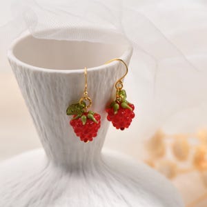 May include: A pair of gold-colored dangle earrings featuring red raspberry charms. Each charm has a textured surface, green leaves, and a gold-colored hook. The earrings are displayed against a white background.