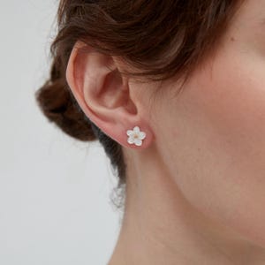 Könnte beinhalten: Nahaufnahme eines Ohrs mit einem weißen, blütenförmigen Ohrstecker. Der Ohrring hat eine kleine goldfarbene Mitte. Der Ohrring ist ein zartes Blumendesign.