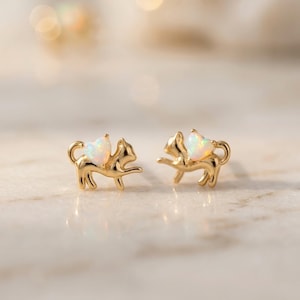 May include: A pair of gold-colored cat-shaped stud earrings. Each earring features a cat with opal-colored wings. The earrings are displayed on a light-colored, marble-like surface. The background is blurred with soft, warm tones.