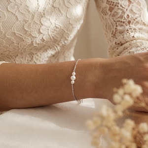 Peut inclure: Un bracelet en argent délicat orné de trois perles blanches. Le bracelet est porté sur un poignet, avec un tissu blanc doux et un vêtement en dentelle en arrière-plan. Les perles sont de tailles variables.