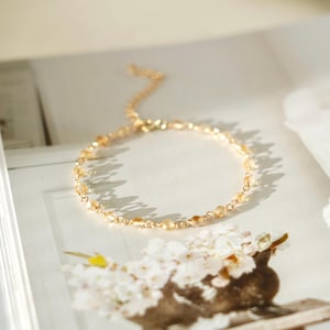 Puede incluir: Una delicada pulsera dorada con pequeñas cuentas de color naranja claro. La pulsera está sobre una superficie blanca, con un arreglo floral en primer plano. La cadena es fina y el cierre es visible.