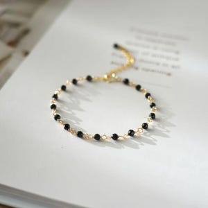 Könnte beinhalten: Ein zartes, goldfarbenes Armband mit kleinen, runden schwarzen Perlen. Die Perlen sind mit einer feinen Goldkette aufgereiht, wodurch ein minimalistisches und elegantes Design entsteht. Das Armband liegt auf einer weißen Oberfläche.