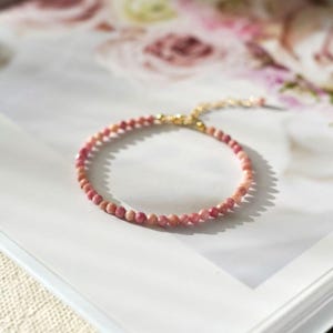 Op de afbeelding: Een delicate armband met kleine, gefacetteerde roze edelstenen. De armband heeft een goudkleurige ketting en sluiting. De edelstenen hebben een zachte roze tint en de armband wordt gepresenteerd op een wit oppervlak.