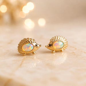Pode incluir: Um par de brincos de tarracha dourados em forma de ouriço. Cada brinco apresenta um corpo de metal texturizado com um pequeno olho preto e um cabochão de opala oval, liso e iridescente, cravado no centro das costas do ouriço.