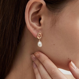 Può includere: Orecchini dorati con un design ad anello irregolare unico. Ogni orecchino presenta una perla bianca a goccia pendente. Gli orecchini sono mostrati su un orecchio, evidenziando il loro stile elegante e minimalista.