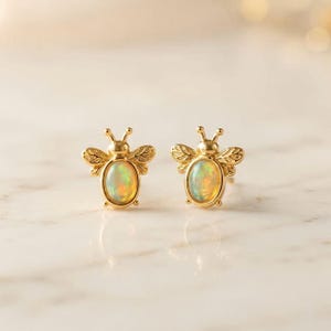 May include: A pair of gold bee-shaped stud earrings. Each earring features a central oval-shaped iridescent opal stone set within a polished gold body, with detailed wing patterns and small antennae extending from the top of the bee design.
