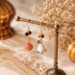 May include: A pair of gold-tone drop earrings featuring mismatched charms. One earring displays an orange jack-o'-lantern, while the other features a white ghost. Both earrings are accented with small, multi-colored faceted beads.