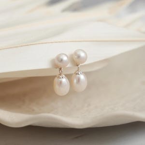 May include: A pair of drop earrings featuring two white pearls each. The top pearl is round and sits directly on the ear, while the bottom pearl is oval-shaped and hangs from a small silver-toned metal cap, creating a delicate dangling effect.