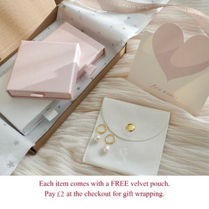 May include: A white velvet pouch with a gold button closure and a small pearl earring inside. The pouch is sitting on a white surface with a pink box and a card that says "Best wishes".