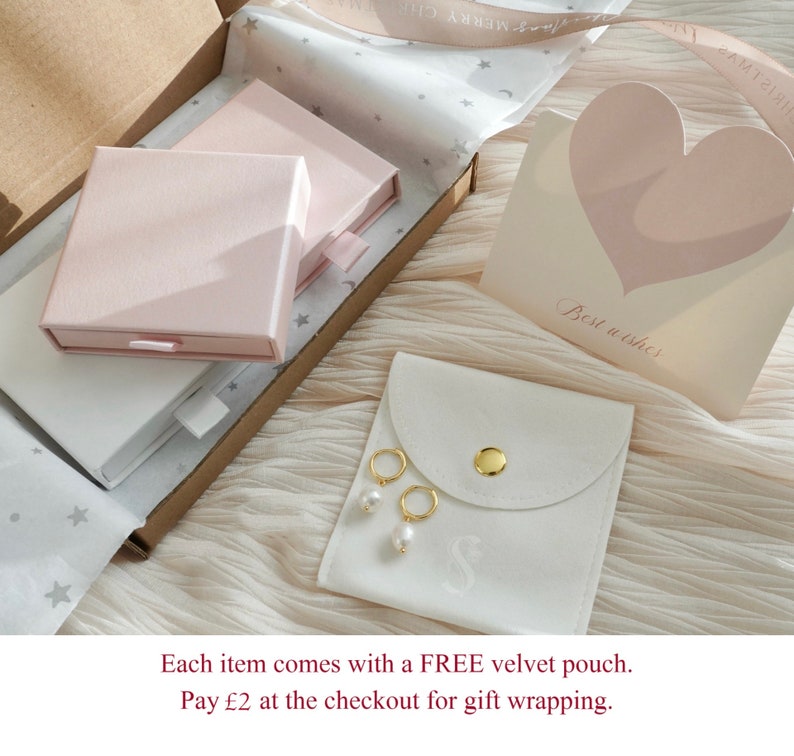 May include: A white velvet pouch with a gold button closure and a small pearl earring inside. The pouch is sitting on a white surface with a pink box and a card that says "Best wishes".