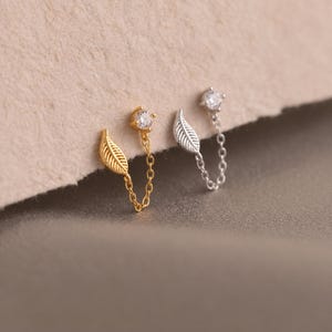 Peut inclure: Deux boucles d'oreilles à double perçage, une dorée et une argentée. Chaque boucle présente un petit strass rond transparent relié par une fine chaîne à une breloque en forme de feuille. Le design permet de relier deux perçages.