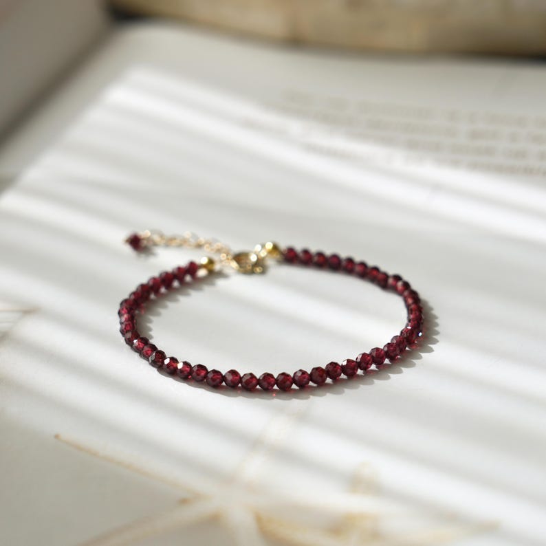 Pu&ograve; includere: Un delicato braccialetto con piccole pietre preziose rosse scuro sfaccettate. Il braccialetto ha una chiusura e una catena color oro ed &egrave; presentato su una superficie bianca. Le pietre preziose sono uniformemente distanziate.