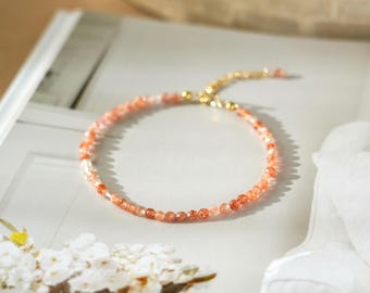 May include: A delicate bracelet composed of small, faceted, translucent orange and peach-colored gemstone beads. The bracelet features a gold-toned metal clasp and an adjustable chain extension, creating a circular shape on a flat surface.