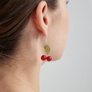 Peut inclure: Boucles d'oreilles pendantes avec un motif de cerise. Chaque boucle d'oreille comporte deux perles de cerise rouges, une feuille verte et un crochet argenté. Les boucles d'oreilles sont présentées sur un fond clair.