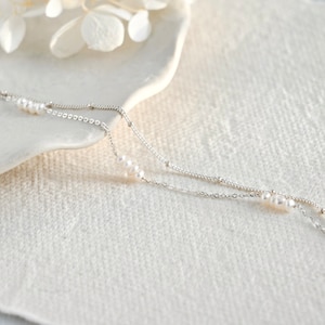 May include: A delicate silver chain necklace with two strands and small white pearls. The necklace is on a white surface.