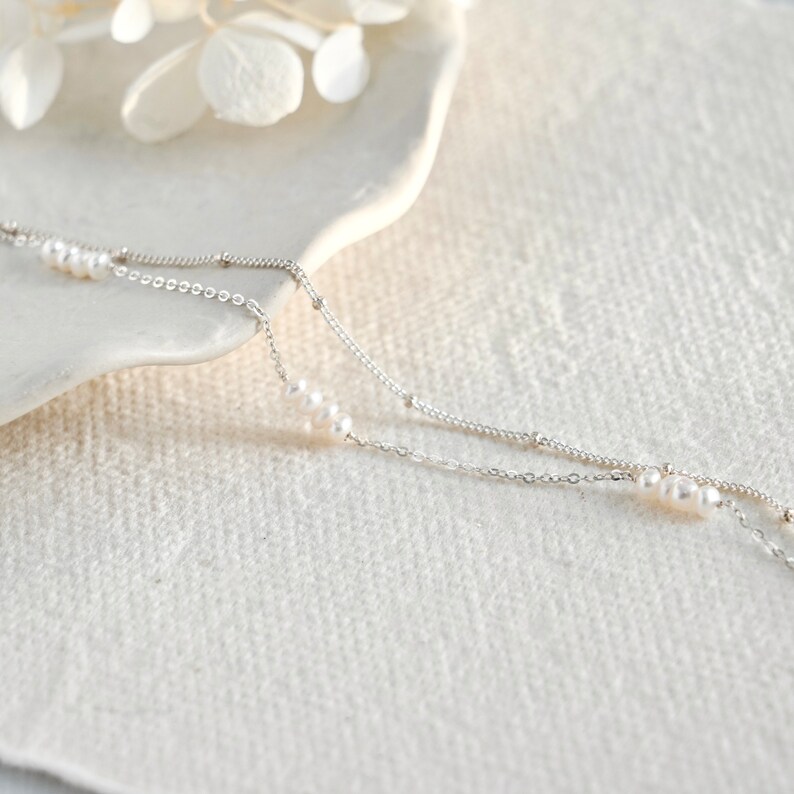 May include: A delicate silver chain necklace with two strands and small white pearls. The necklace is on a white surface.