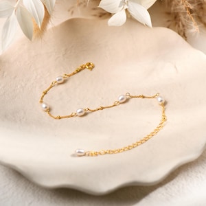 May include: A delicate gold-colored bracelet featuring small, round pearls. The bracelet has a fine chain with small, dotted accents and a clasp closure. The pearls are evenly spaced along the chain, creating a classic and elegant look.