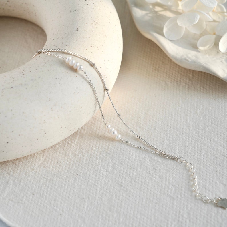 May include: A delicate silver chain bracelet with small white pearls. The bracelet has two chains, one with a delicate chain and the other with a slightly thicker chain.