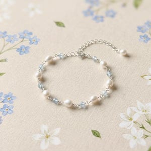 Könnte beinhalten: Ein zartes Armband mit weißen Perlen und hellblauen Perlen, die auf einer silbernen Kette aufgereiht sind. Das Armband wird auf einer cremefarbenen Oberfläche mit floralen Akzenten präsentiert und zeigt ein klassisches Schmuckdesign.