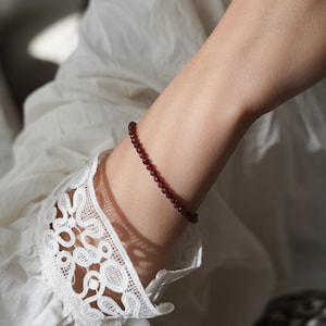Pu&ograve; includere: Un delicato braccialetto composto da piccole perline rosse scure sfaccettate. Il braccialetto &egrave; indossato su un polso, con una manica di pizzo bianco sullo sfondo. Le perline sembrano essere di dimensioni uniformi e infilate.