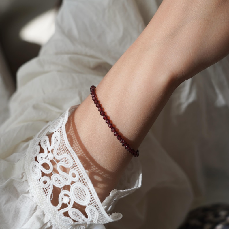 Pu&ograve; includere: Un delicato braccialetto composto da piccole perline rosse scure sfaccettate. Il braccialetto &egrave; indossato su un polso, con una manica di pizzo bianco sullo sfondo. Le perline sembrano essere di dimensioni uniformi e infilate.