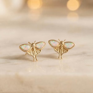 May include: A pair of gold stud earrings shaped like moths, featuring iridescent white opal inlays on the wings. The metal has a polished finish with detailed line work on the lower wing sections, creating a symmetrical insect design.