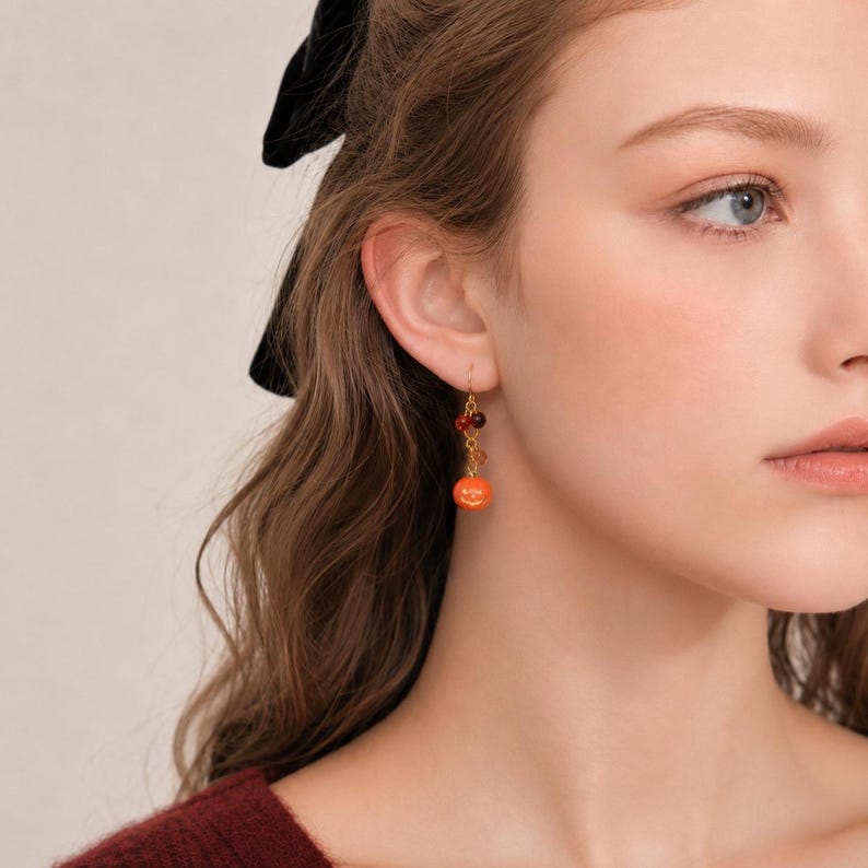 May include: A gold-toned dangle earring featuring a small, round, bright orange pumpkin charm suspended from a delicate chain. The earring includes two smaller, dark-colored beads positioned above the pumpkin charm, attached to a simple hook wire.