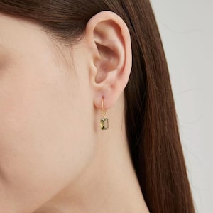 Puede incluir: Un pendiente dorado con una gema rectangular de color verde oliva. El pendiente cuelga de la oreja. La gema es de color verde claro. El pendiente tiene un dise&ntilde;o sencillo y elegante.
