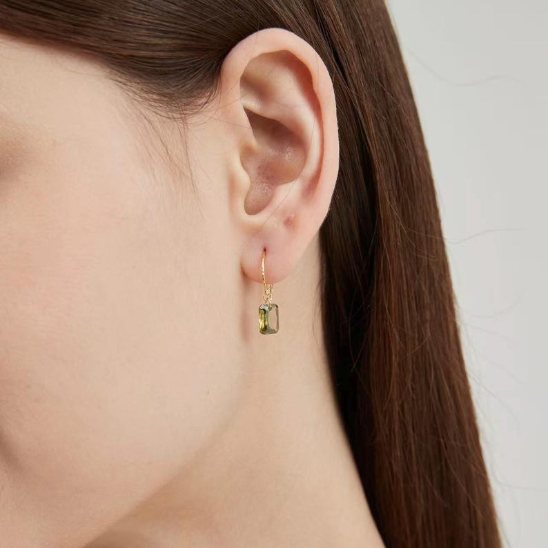 Puede incluir: Un pendiente dorado con una gema rectangular de color verde oliva. El pendiente cuelga de la oreja. La gema es de color verde claro. El pendiente tiene un dise&ntilde;o sencillo y elegante.