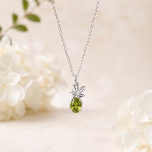 May include: A silver necklace featuring an oval-cut lime green peridot gemstone pendant. The pendant is accented with a small clear round gemstone and a silver leaf-shaped design element. The pendant hangs from a delicate silver chain.