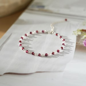 Könnte beinhalten: Ein filigranes Silberarmband mit kleinen, runden, tiefroten Edelsteinen. Das Armband ist an einer feinen Silberkette aufgereiht und liegt auf einer weißen Oberfläche, mit weicher Beleuchtung und einem verschwommenen Hintergrund.