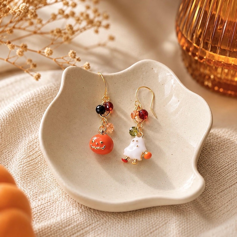 May include: A pair of gold-tone drop earrings featuring charms of an orange jack-o'-lantern and a white ghost. Each earring is accented with small, multi-colored faceted beads in shades of red, black, green, and orange on gold-tone hooks.