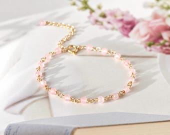 Könnte beinhalten: Ein zartes, goldfarbenes Kettenarmband mit kleinen, runden, blassrosa Perlen, die gleichmäßig entlang der Glieder verteilt sind. Das Armband verfügt über einen runden Verschluss und eine verstellbare Verlängerungskette.