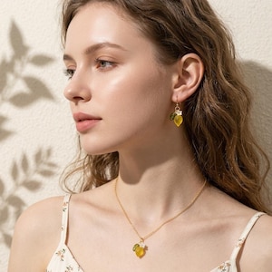 May include: A gold necklace and matching earrings featuring a lemon design. The pendant and earrings have a yellow lemon shape with green leaves and a small white flower. The jewelry is worn by a person with long wavy hair.
