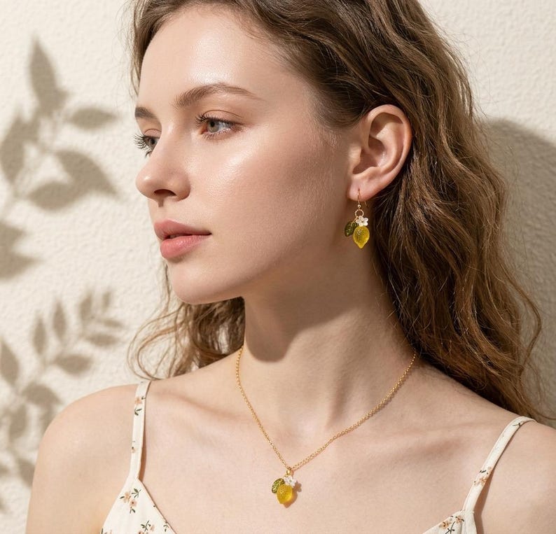 May include: A gold necklace and matching earrings featuring a lemon design. The pendant and earrings have a yellow lemon shape with green leaves and a small white flower. The jewelry is worn by a person with long wavy hair.