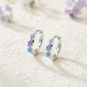 May include: A pair of silver hoop earrings featuring small, enamel-coated flowers in shades of light blue, purple, and white. Each hoop is decorated with three distinct floral accents, showcasing a delicate design with metallic silver edges.