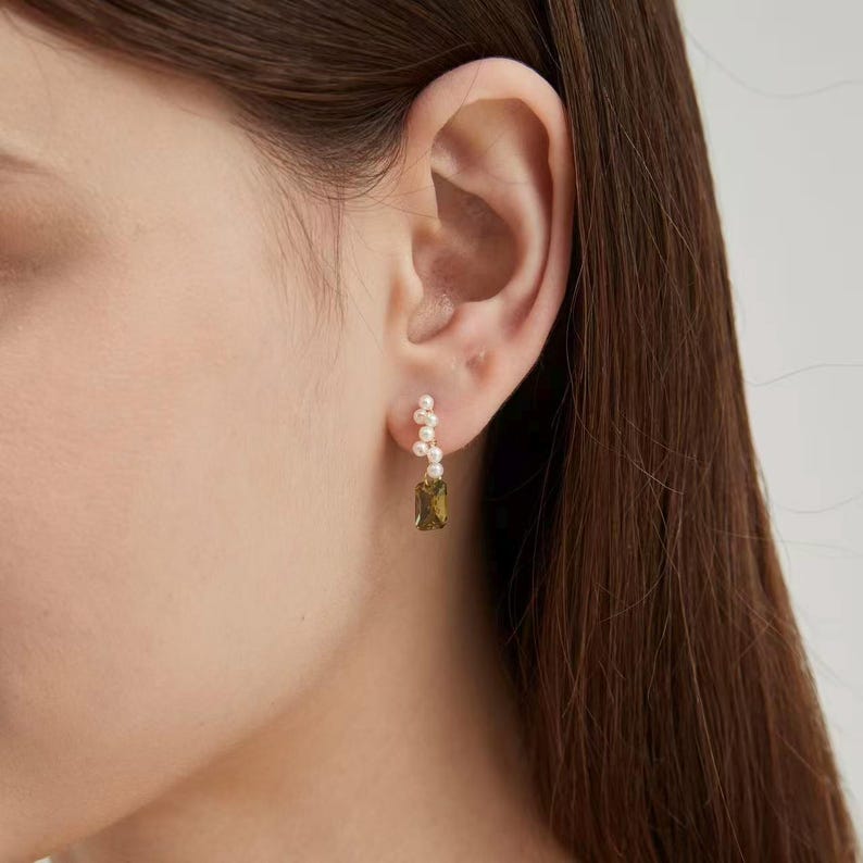 Puede incluir: Un pendiente dorado con un grupo de peque&ntilde;as perlas blancas y una gema rectangular de color verde oliva. El pendiente se muestra en una oreja, mostrando su delicado dise&ntilde;o y contraste de color.