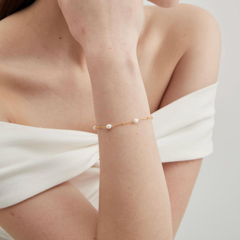 May include: A delicate gold bracelet featuring five evenly spaced, lustrous white pearls. The bracelet is worn on a wrist, showcasing its simple elegance. The gold chain has a subtle texture, adding to the bracelet's refined appearance.