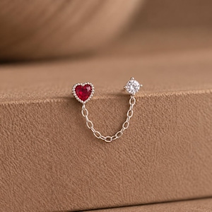 May include: A silver double-stud earring connected by a delicate chain. One stud features a red heart-shaped gemstone with a beaded border, while the other stud is a round, clear, faceted gemstone set in a four-prong silver mount.