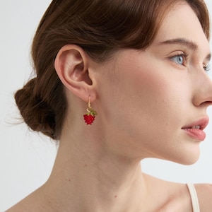 May include: A gold-colored earring with a red, textured raspberry design and green leaves. The earring is hanging from a gold-colored hook. The earring is a piece of jewelry.