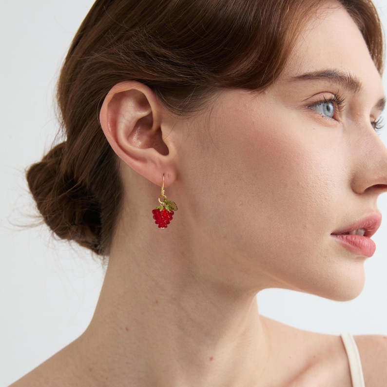 May include: A gold-colored earring with a red, textured raspberry design and green leaves. The earring is hanging from a gold-colored hook. The earring is a piece of jewelry.