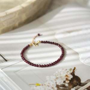 Pu&ograve; includere: Un delicato braccialetto con piccole perline sfaccettate di colore rosso intenso. Il braccialetto ha una chiusura e una catena regolabile color oro. Il gioiello &egrave; esposto su una superficie bianca con un'illuminazione soffusa, suggerendo eleganza e semplicit&agrave;.