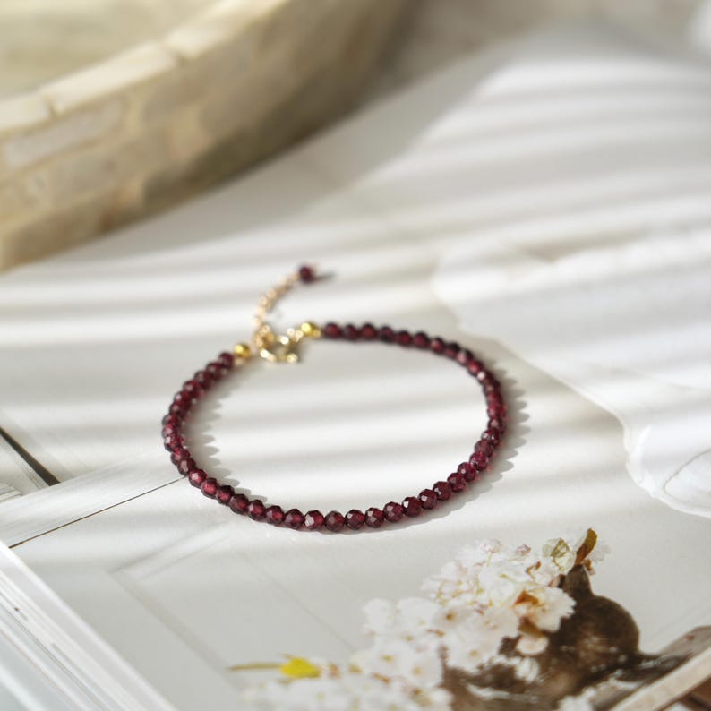 Pu&ograve; includere: Un delicato braccialetto con piccole perline sfaccettate di colore rosso intenso. Il braccialetto ha una chiusura e una catena regolabile color oro. Il gioiello &egrave; esposto su una superficie bianca con un'illuminazione soffusa, suggerendo eleganza e semplicit&agrave;.