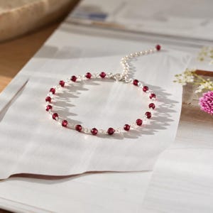 Op de afbeelding: Een delicate armband met kleine, gefacetteerde donkerrode granaatkralen afgewisseld met dunne zilverkleurige metalen schakels. Het cirkelvormige sieraad bevat een korte verlengketting met een enkele kraal aan het uiteinde.