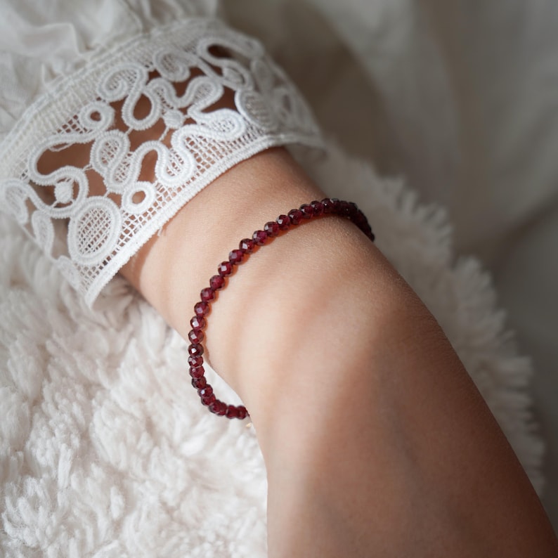 Pu&ograve; includere: Un delicato braccialetto con piccole perline rotonde, rosso scuro. Il braccialetto &egrave; indossato su un polso, con una manica di pizzo bianco sullo sfondo. Le perline sembrano essere di dimensioni uniformi e infilate insieme.