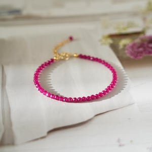 May include: A delicate, magenta-colored beaded bracelet with a gold-toned clasp. The bracelet is composed of small, round beads and is displayed on a white surface. The bracelet is a piece of jewelry.