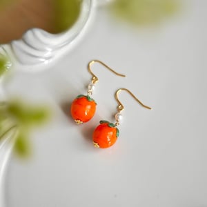 Könnte beinhalten: Ein Paar orangefarbene Kaki-Ohrringe mit goldfarbenen Haken und grünen Blättern. Jeder Ohrring hat eine kleine weiße Perle und goldfarbene Akzente. Die Ohrringe sind vor einem weißen Hintergrund platziert.