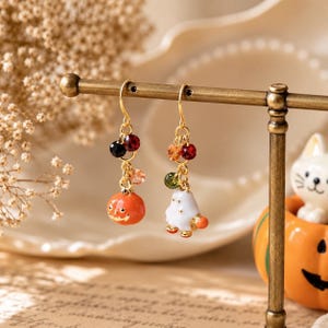 May include: A pair of gold-tone drop earrings featuring dangling charms. One earring displays an orange pumpkin with a carved face, while the other shows a white ghost figure. Both are accented with small, multi-colored faceted beads.