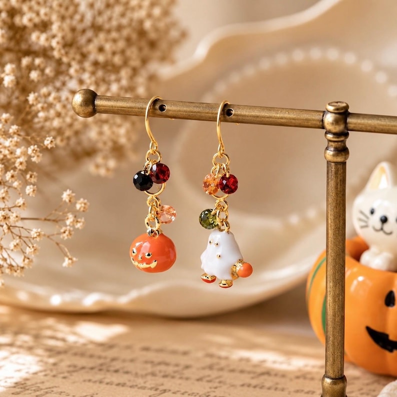 May include: A pair of gold-tone drop earrings featuring dangling charms. One earring displays an orange pumpkin with a carved face, while the other shows a white ghost figure. Both are accented with small, multi-colored faceted beads.