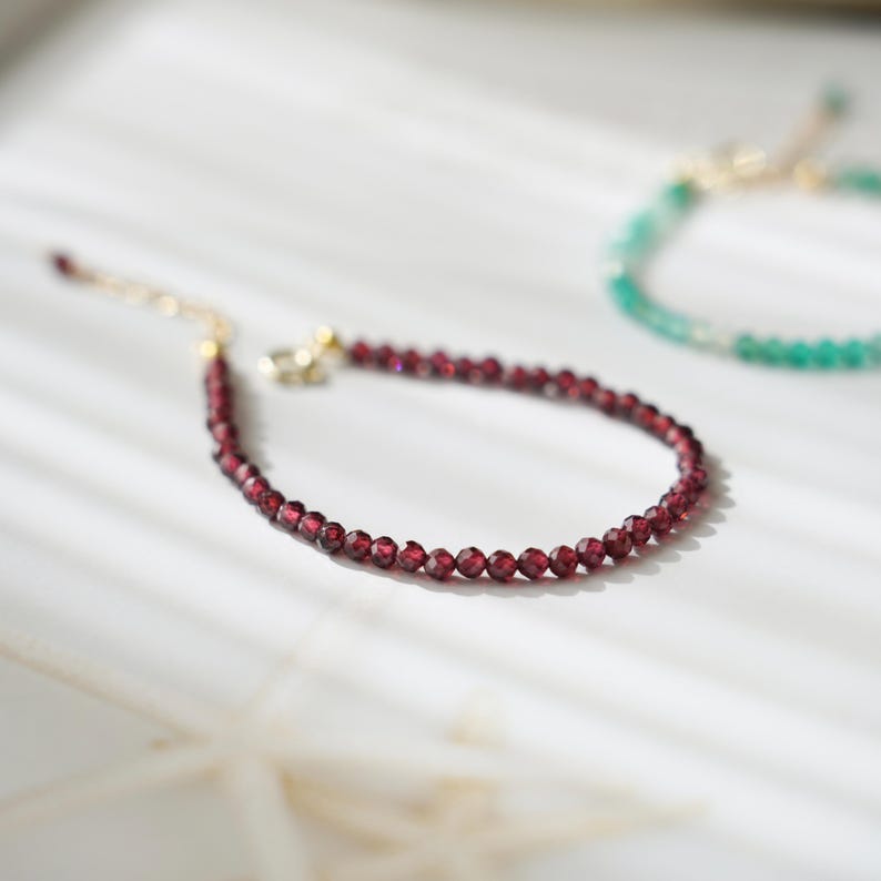 Pu&ograve; includere: Un delicato braccialetto con piccole perline sfaccettate di colore rosso intenso. Il braccialetto ha una catena e una chiusura color oro. Un altro braccialetto con perline verdi &egrave; leggermente sfocato sullo sfondo.